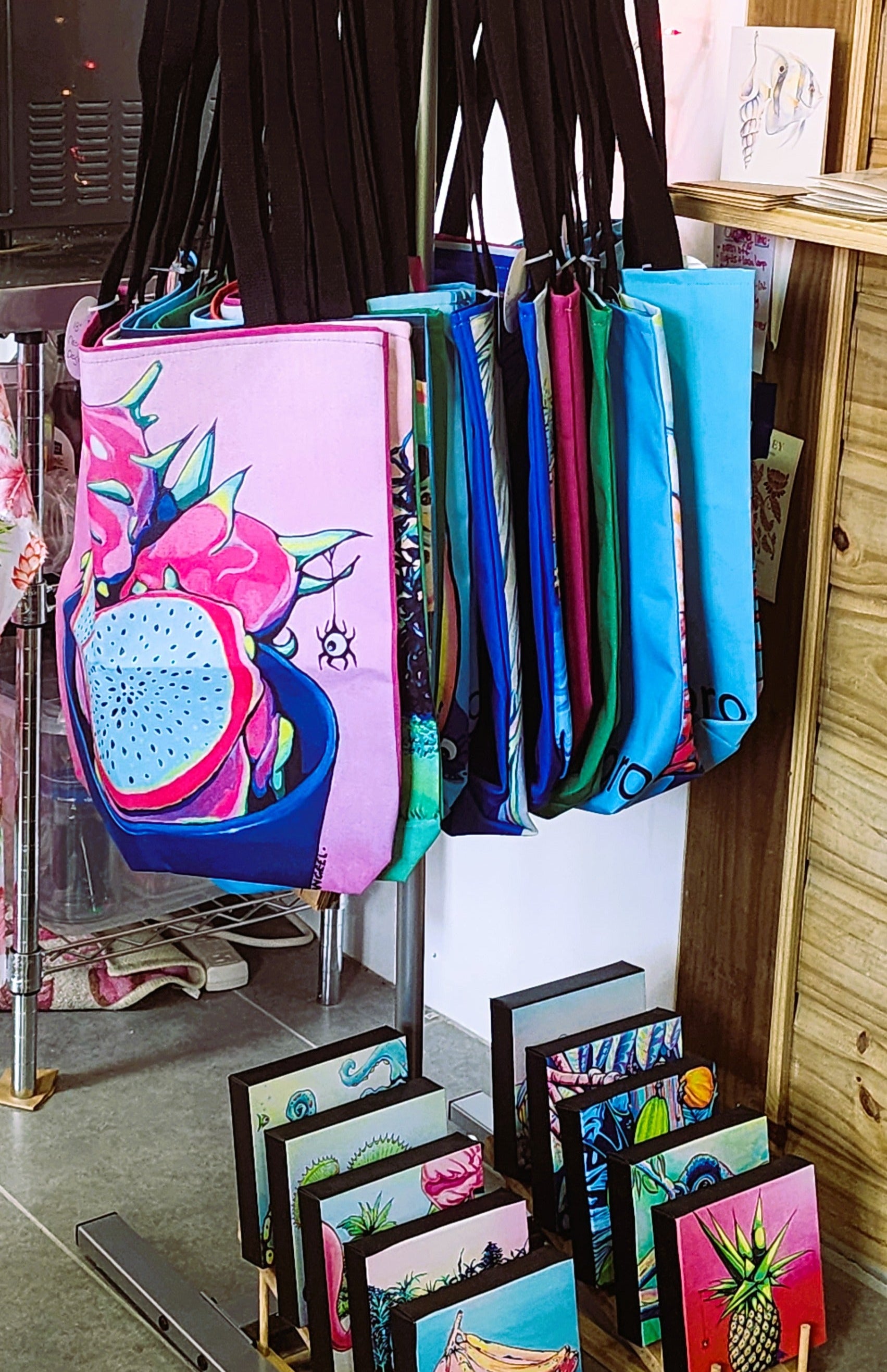 A display of colorful tote bags hanging on a stand in a retail shop, alongside small framed art prints on wooden racks. The bags feature vibrant tropical and floral designs, including pink, blue, green, and black patterns.