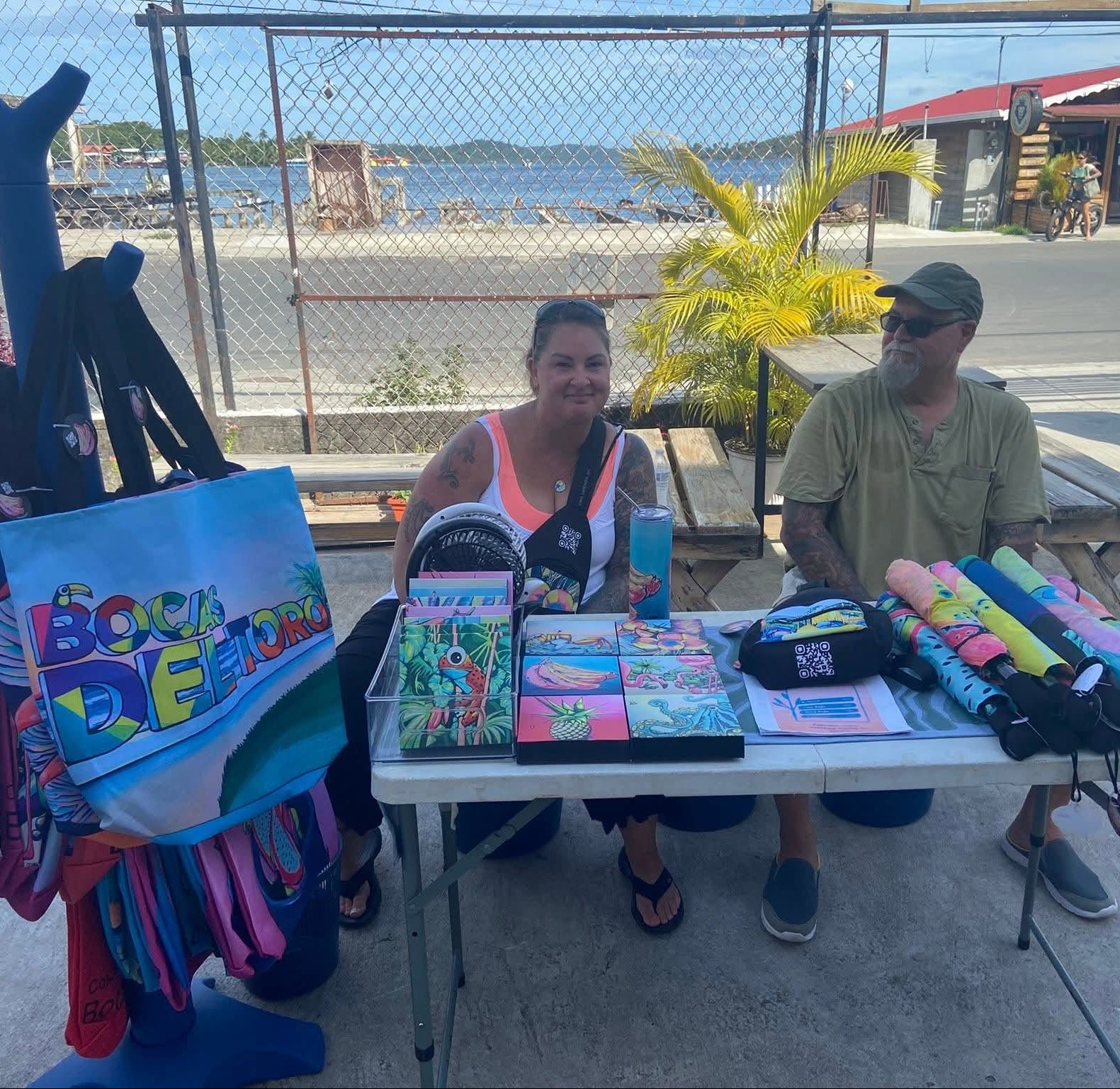Two people sitting behind a table with bags and accessories near a beach.
