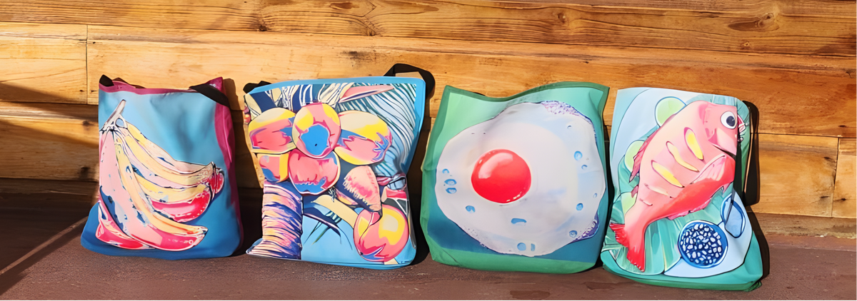 Four colorful beach tote bags with tropical and food-themed prints are lined up against a wooden wall on a concrete floor, with a pair of sandals nearby.